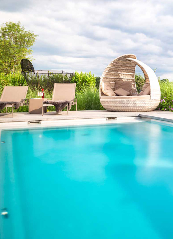 Türkisfarbener Pool vor grün bepflanztem Garten mit modernen Lounge-Sesseln und gemütlicher Korbkugel, passend zu Goroncy Referenzen.