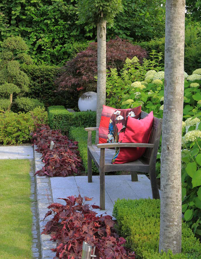 Bei Goroncy Referenzen: Eine Sitzbank mit roten Kissen im grünen Garten, umgeben von gepflastertem Weg und dunkelrotem Bodendecker.