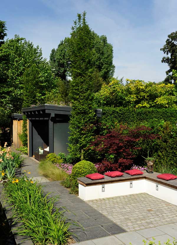Ruhiger grüner Garten mit gepflastertem Weg, dunklem Sichtschutz und roten Kissen am Sitzbereich, umgeben von dichtem Hecken- und Beetgrün.
